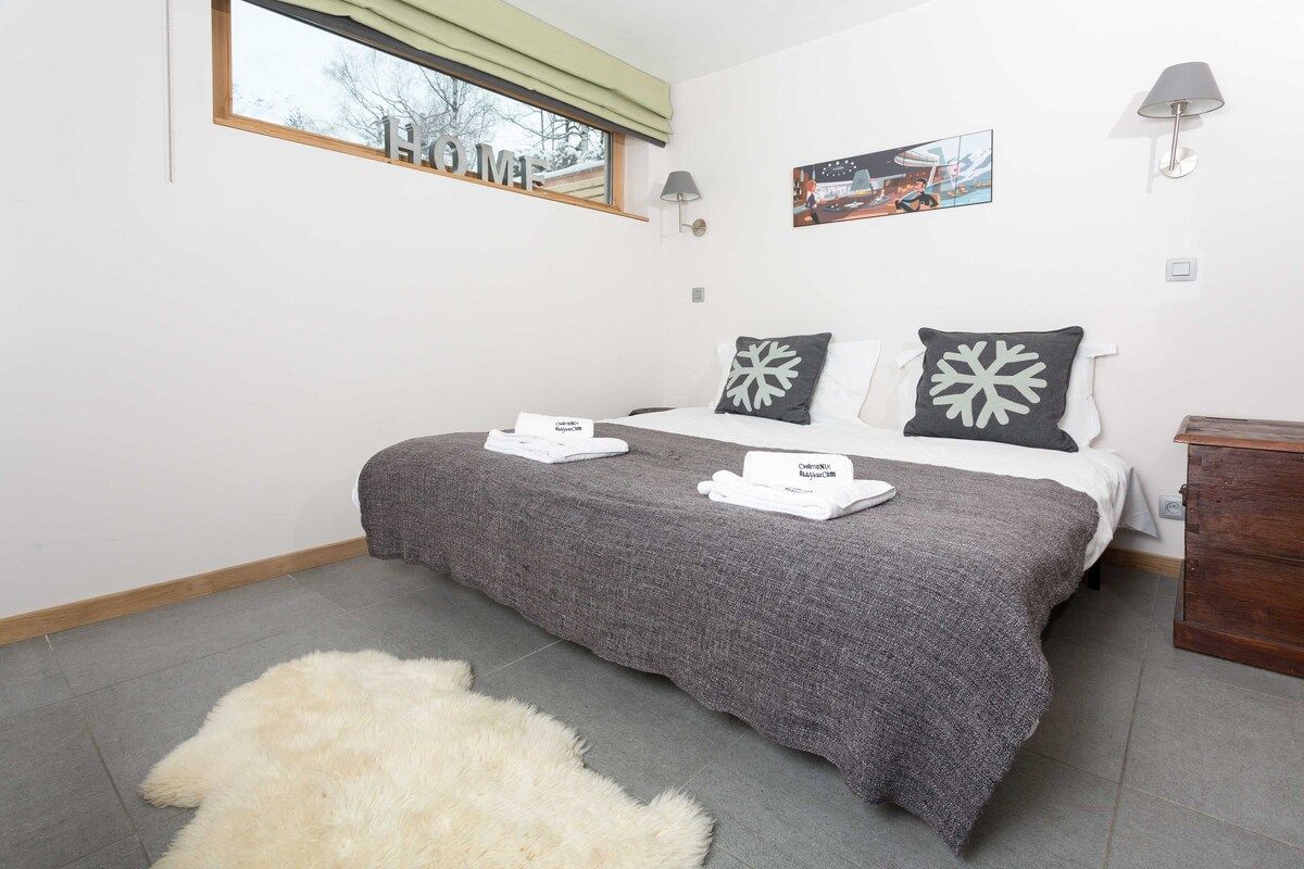 Un dormitorio con cama, almohadas, una mesita de noche, lámpara, ventana con vistas y alfombra.