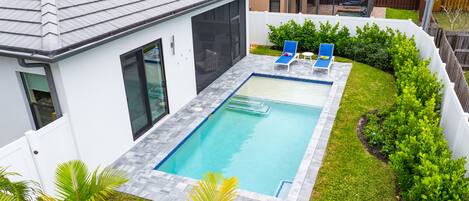 A rectangular swimming pool with a tiled deck, two lounge chairs, and a small garden area with plants.