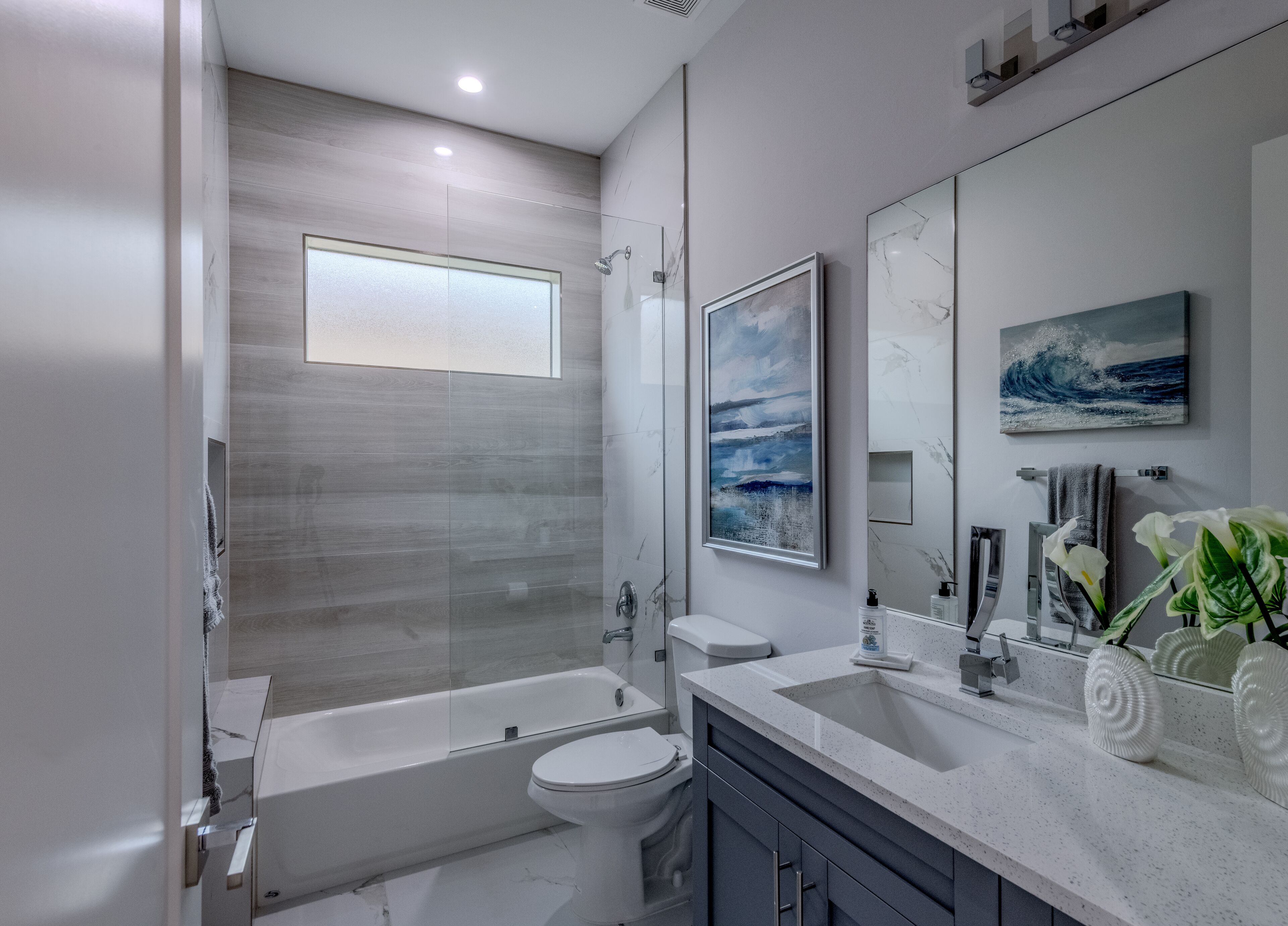 Une salle de bain moderne avec une baignoire, des toilettes et un lavabo. Deux œuvres d’art encadrées ornent le mur, et une fenêtre se trouve au-dessus de la baignoire.