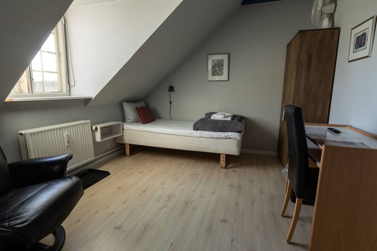 Ein kleines Schlafzimmer im Loft-Stil mit einem Einzelbett, einem Schreibtisch und einem Sessel. Unter dem Fenster befindet sich eine Heizung, ein schwarzer Sessel und ein Holzschrank.