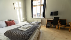 A neatly made bed with pillows and a blanket, a desk with a chair, a television, and a window with curtains. - Hotel Rønne H (Ronne)
