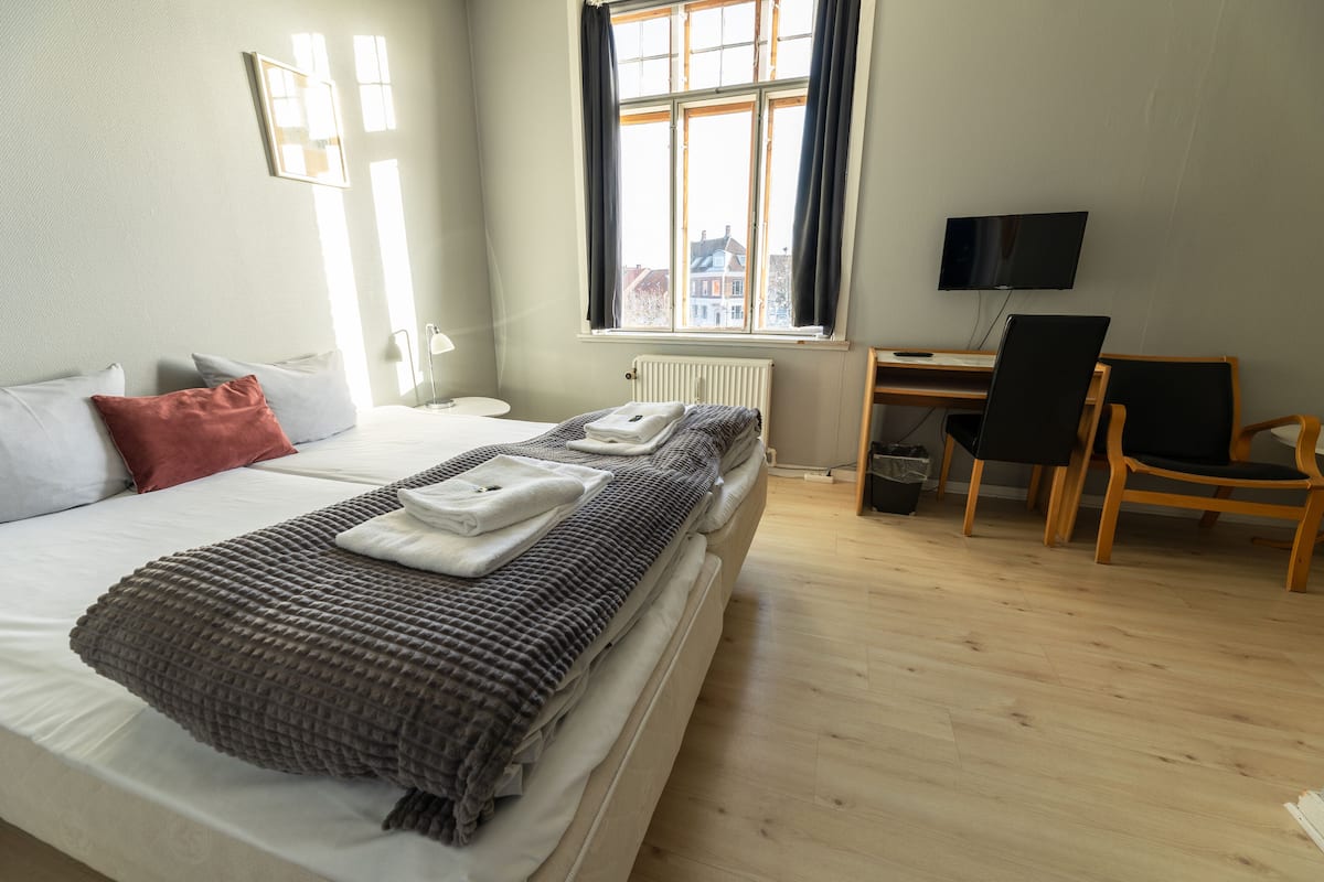 Ein ordentlich bezogenes Bett mit Kissen und Decke, ein Schreibtisch mit Stuhl, ein Fernseher und ein Fenster mit Vorhängen.
