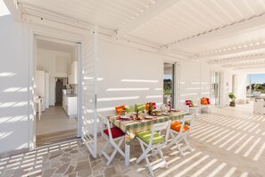 Uma área de refeições ao ar livre com uma mesa posta para quatro pessoas, rodeada por cadeiras brancas com almofadas coloridas.