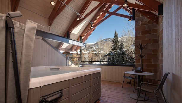 A wooden deck with a hot tub, a table with chairs, and a view of snow-covered mountains.