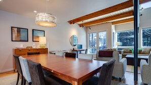 A modern dining area with a wooden table and chairs, a chandelier, and a view of the outdoors through large windows.