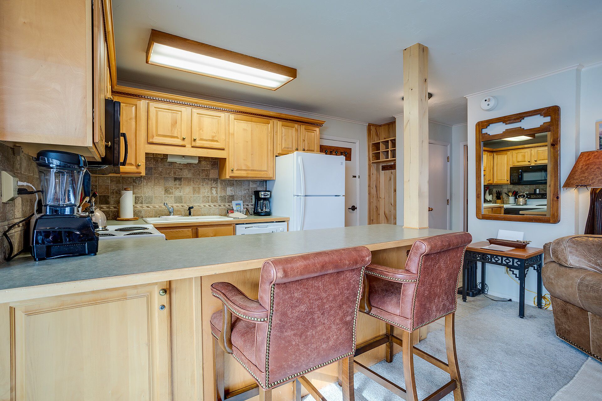 A kitchen with wooden cabinets, a refrigerator, a coffee maker, and a blender. There are two bar stools and a small table with a lamp.