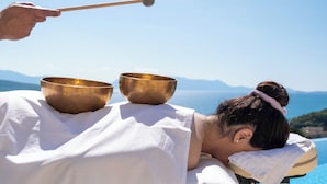 Une personne reçoit un massage avec deux bols dorés placés sur son dos, le tout se déroulant dans un cadre pittoresque, avec une vue sur la montagne et la mer.