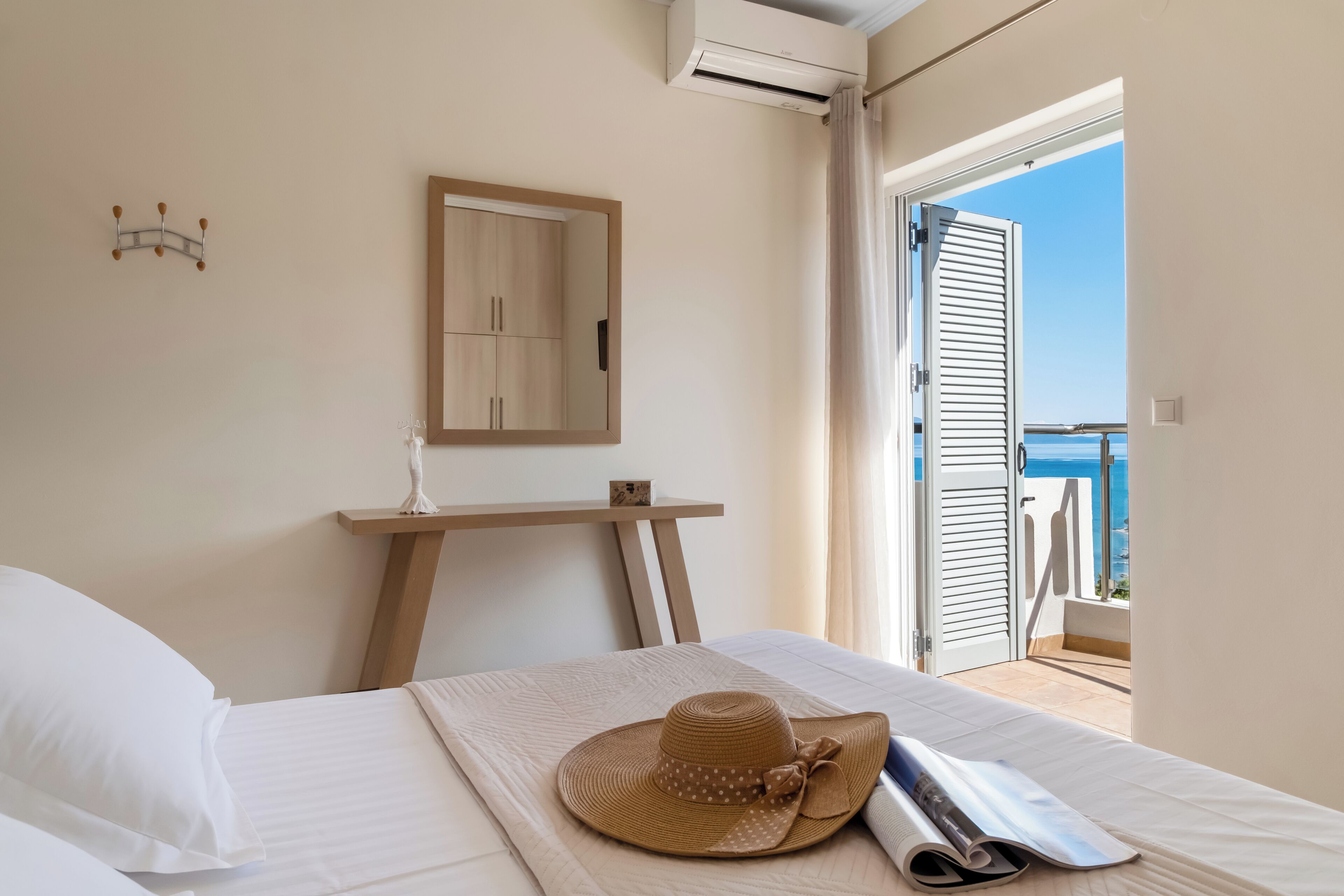 A hotel room with a bed, a small desk, a mirror, and a view of the sea through an open door.