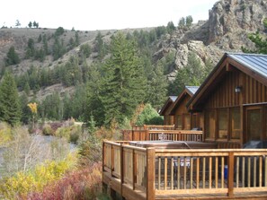 A wooden cabin with a deck, surrounded by a forest and a rocky hillside. - Beautiful 2 Bedroom Cabin on Taylor River with Private Hot Tub (Almont)