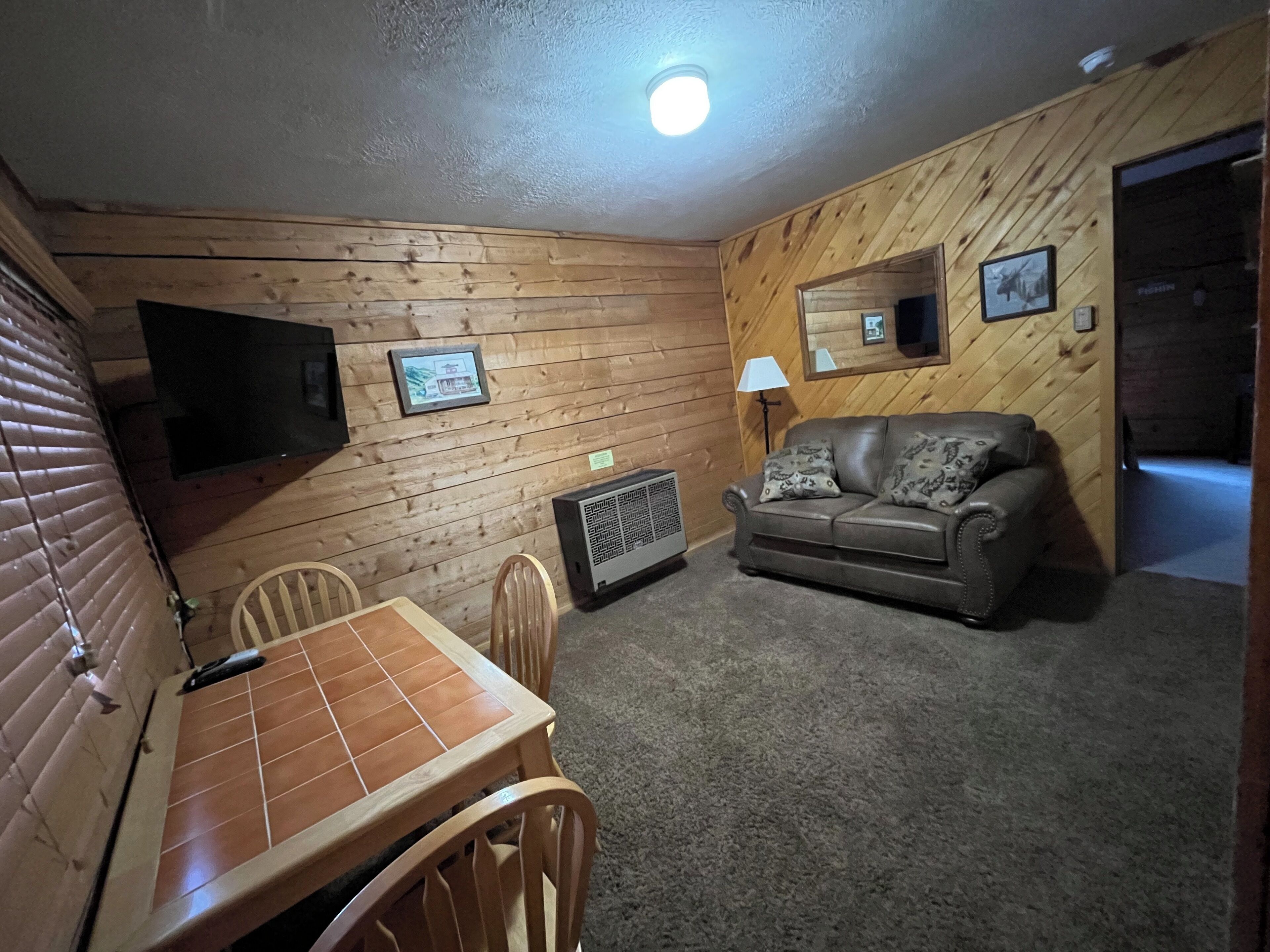 A room with wooden walls, a leather sofa, a TV mounted on the wall, a table with chairs, and a heater.