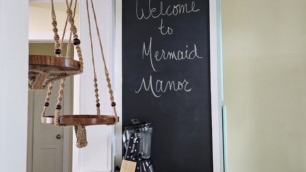 Eine Küche mit einem Herd, hängenden Holzdekorationen und einer Tafel mit der Aufschrift 'Welcome to Mermaid Manor'.