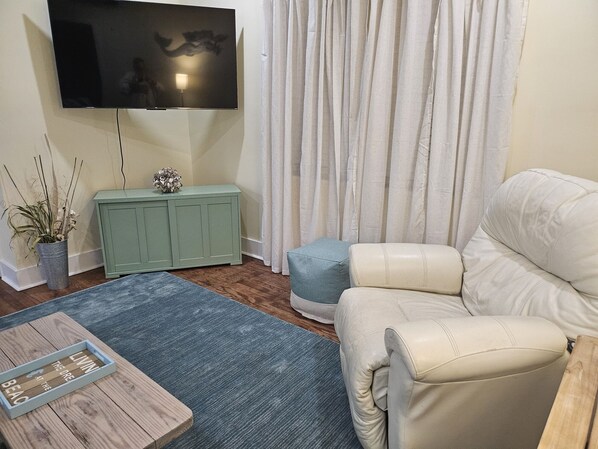 Ein Wohnzimmer mit einem an der Wand montierten Flachbildfernseher, einem grünen Schrank, einem weißen Sofa, einem blauen Hocker und einem Holztisch mit einer Schale dekorativer Gegenstände.