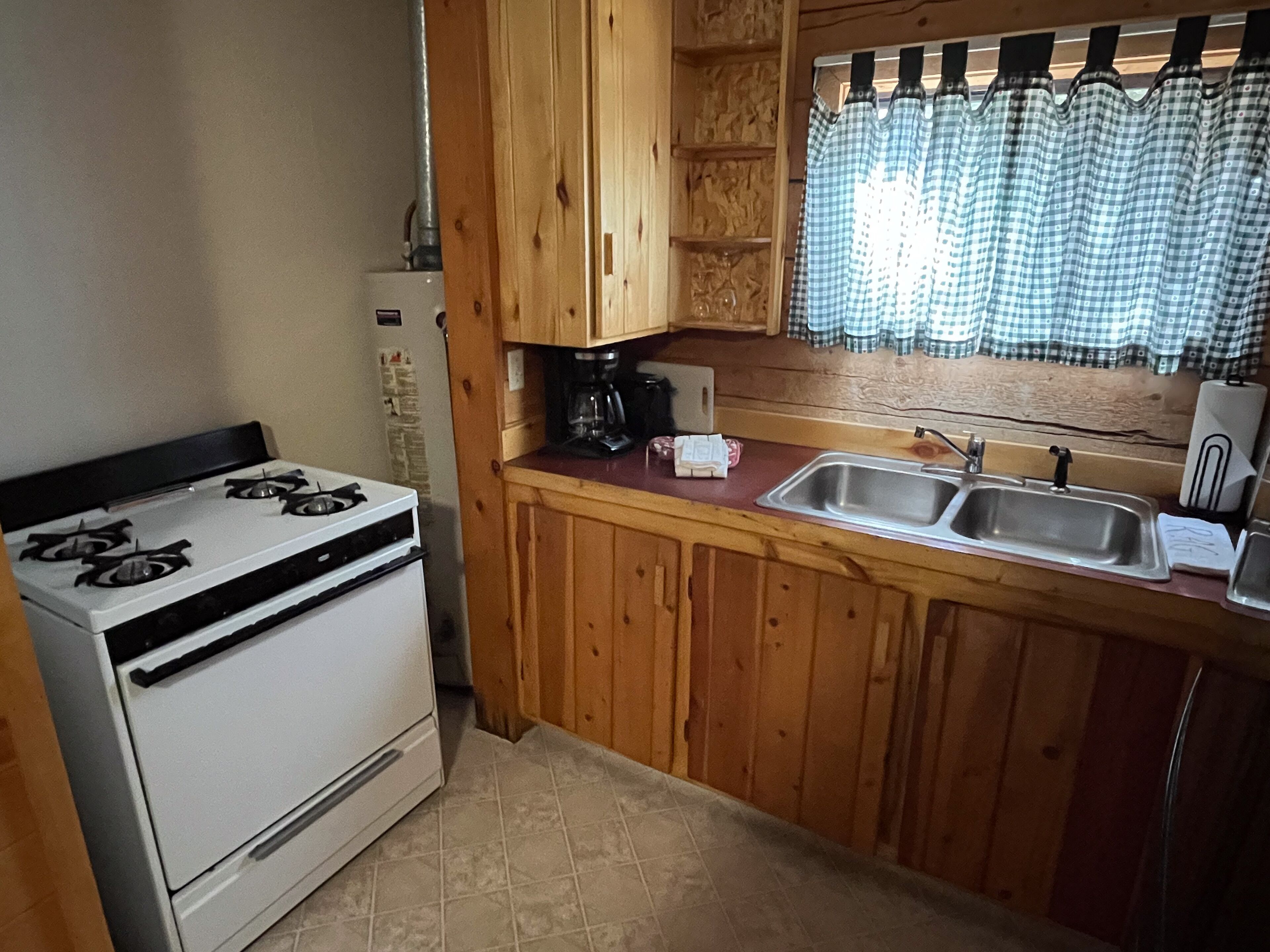 A compact kitchen with wooden cabinets, a gas stove, a sink, and a window with patterned curtains.