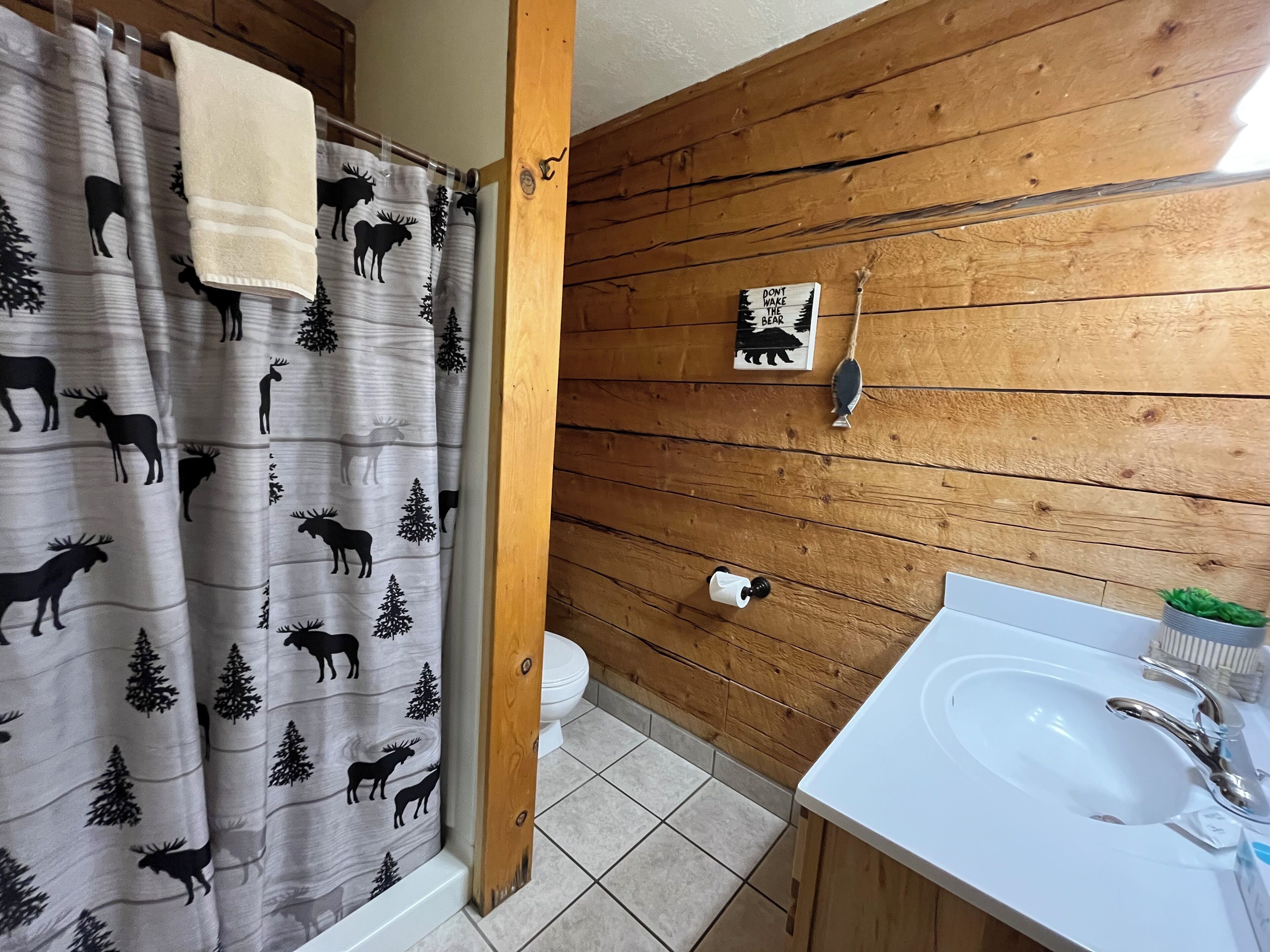 Un baño con pared de madera, cortina de ducha con motivos de alces y pinos, inodoro y lavabo con espejo.