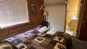Un dormitorio con pared de madera, colcha acolchada, mesita de noche con lámpara y ventana con persianas.