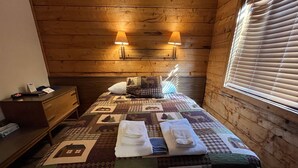 Une chambre confortable avec une tête de lit en bois, deux lampes fixées au mur, une couvre-lit matelassé, une table de chevet avec une lampe et une fenêtre avec des stores.