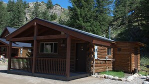 Une cabane en rondins avec un porche, un petit bâtiment attenant et une montagne rocheuse en arrière-plan.
