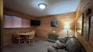 L’intérieur d’un chalet accueillant, avec un revêtement mural en bois, une table à manger entourée de chaises, un canapé, un téléviseur fixé au mur et un luminaire au plafond.