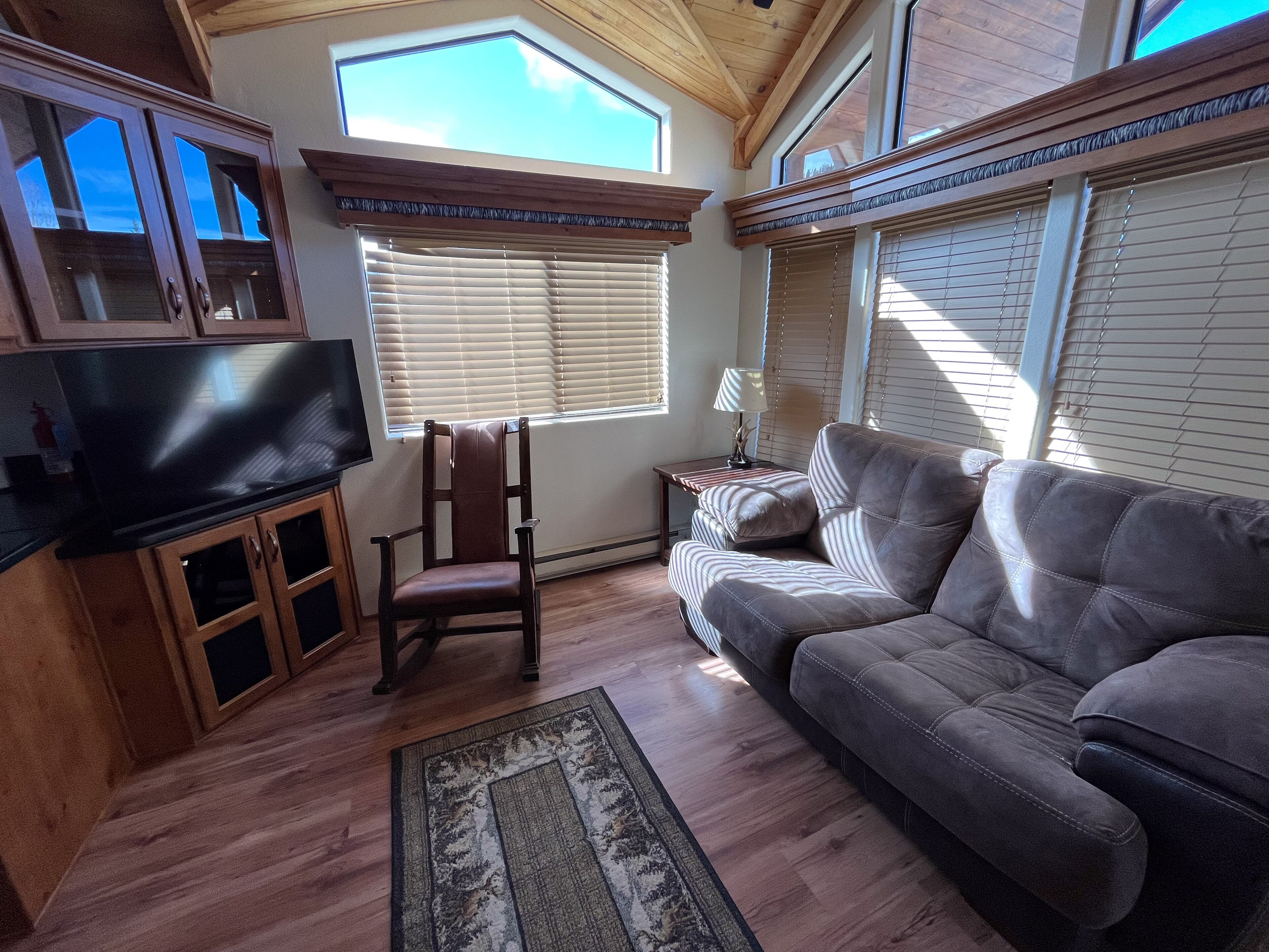 A living room with a wooden floor, a sofa, a chair, a TV, and a window with blinds.