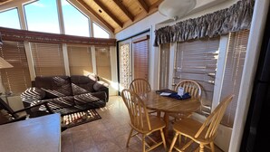 Una sala de estar acogedora con un sofá, una mesa de comedor de madera y sillas. Se ven ventanas amplias con persianas y un ventilador de techo.