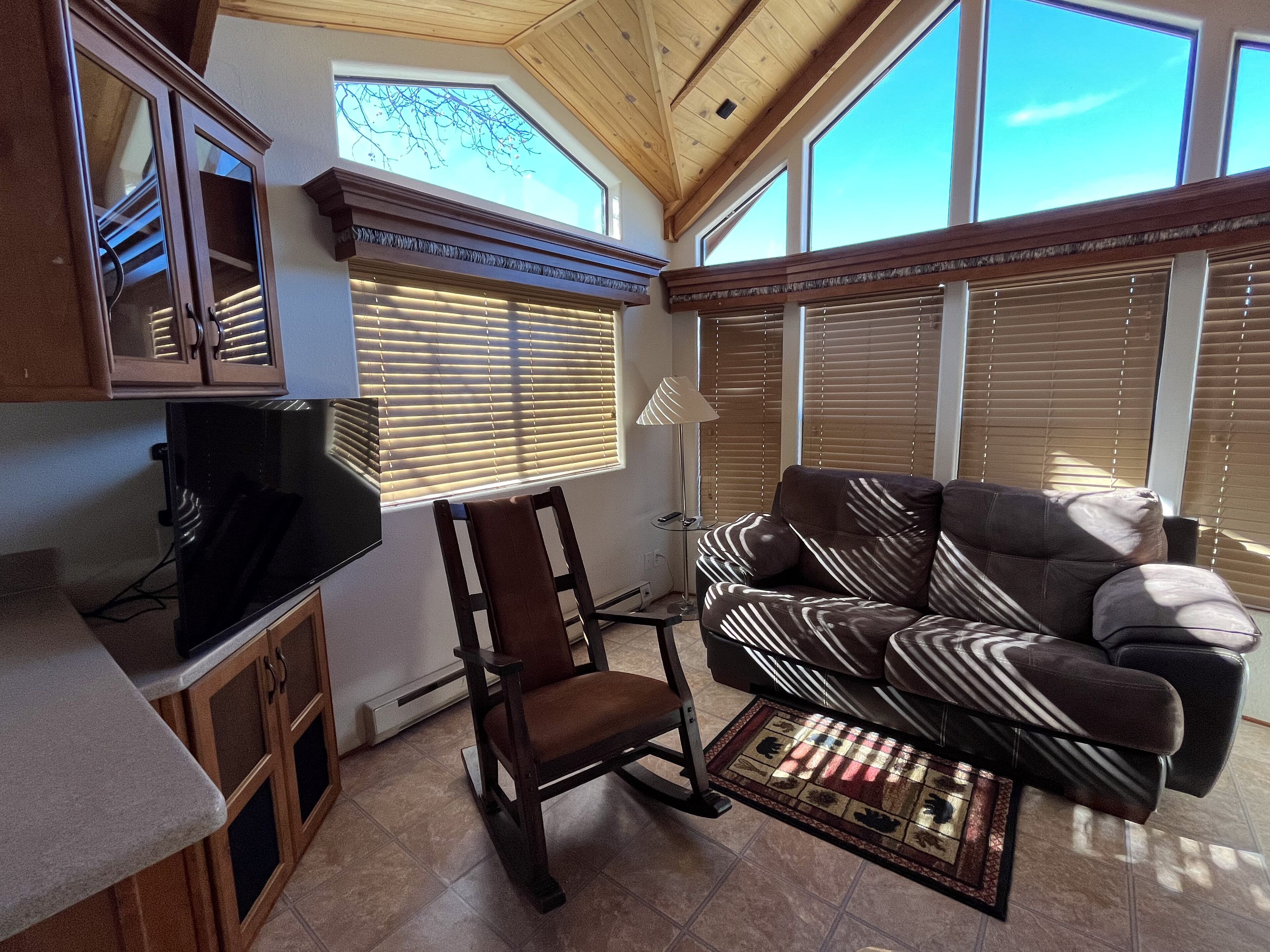 A cozy living room with a brown leather sofa, a wooden chair, and a television on a cabinet. Large windows with blinds allow natural light.