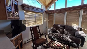 Una sala de estar acogedora con un sofá de cuero marrón, una silla de madera y un televisor sobre un mueble bajo. Amplios ventanales con persianas permiten la entrada de luz natural.