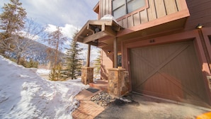 Un bâtiment en bois avec une entrée en pierre, un garage et un paysage enneigé parsemé d’arbres.