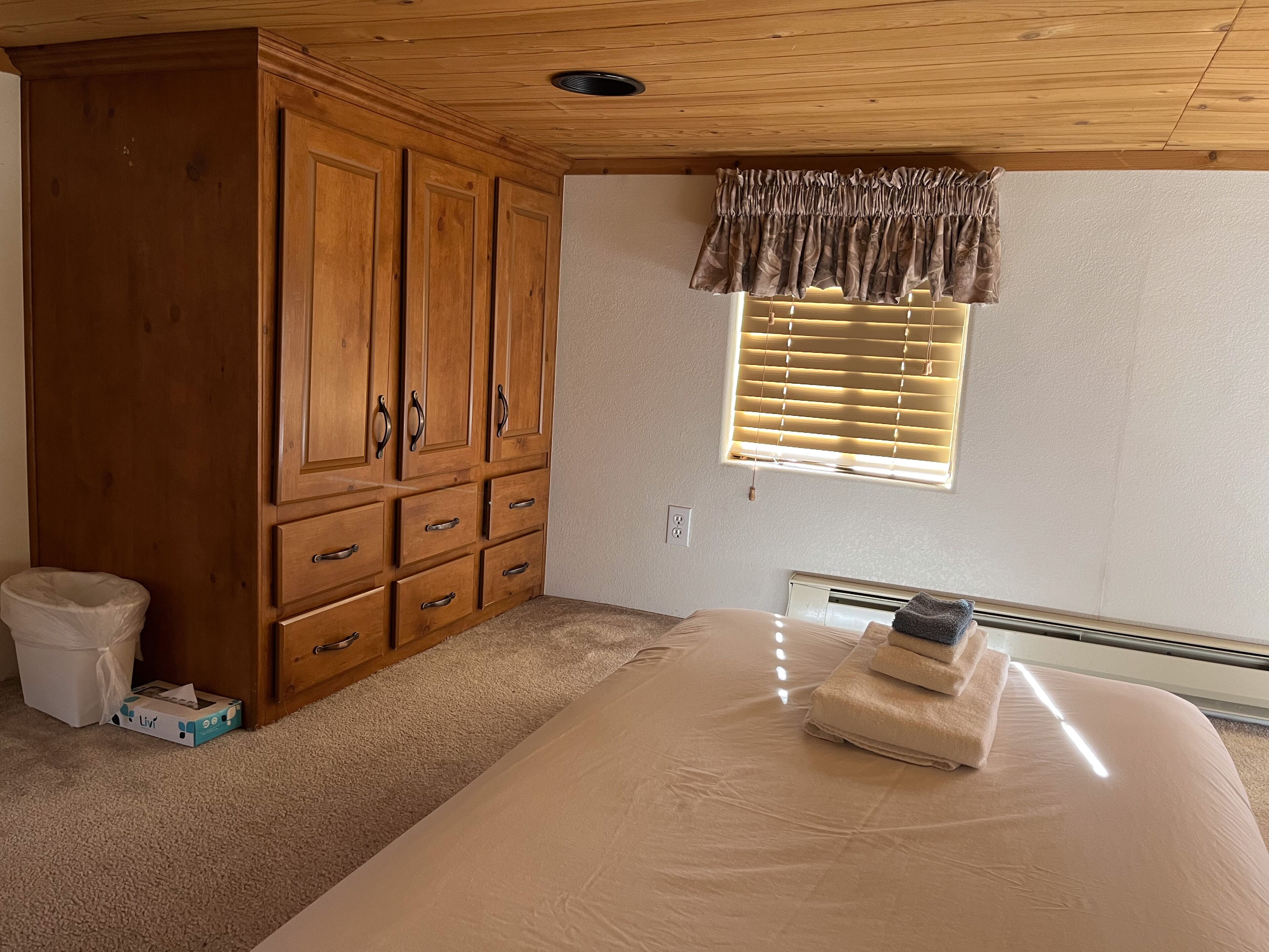 Een slaapkamer met een grote houten garderobe, een enkel bed met een beige matras, een raam met zonwering en een witte prullenbak.