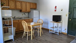 A compact kitchen with wooden cabinets, a white refrigerator, a dining table with chairs, a small TV on a stand, and a fire extinguisher on the wall. - Hotel Style Lodge Room w/Kitchenette (Almont)