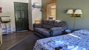 A room with a grey sofa, a bed with floral bedding, a desk with a computer, a wall-mounted lamp, and a door with a fire extinguisher. - Hotel Style Lodge Room w/Kitchenette (Almont)