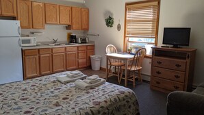 A compact kitchen with wooden cabinets, a microwave, and a sink. There's a dining table with chairs, a TV on a wooden cabinet, and a bed with a floral bedspread. - Hotel Style Lodge Room w/Kitchenette (Almont)