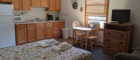 A compact kitchen with wooden cabinets, a microwave, and a sink. There's a dining table with chairs, a TV on a wooden cabinet, and a bed with a floral bedspread.