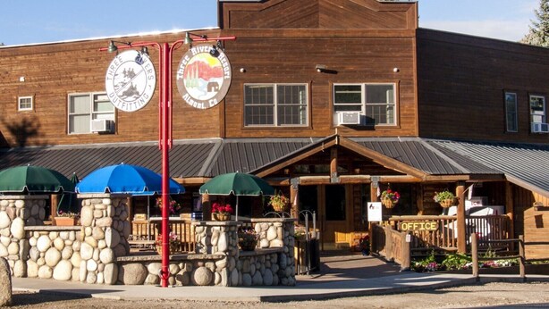 A rustic wooden building with a stone facade, outdoor seating with umbrellas, and a sign for 'Three Rivers Outfitters Co.' - Hotel Style Lodge Room w/Kitchenette (Almont)