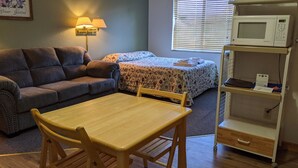 A small, cozy room with a wooden table and chairs, a sofa, a bed with a floral bedspread, a lamp, and a microwave on a shelf. - Hotel Style Lodge Room w/Kitchenette (Almont)