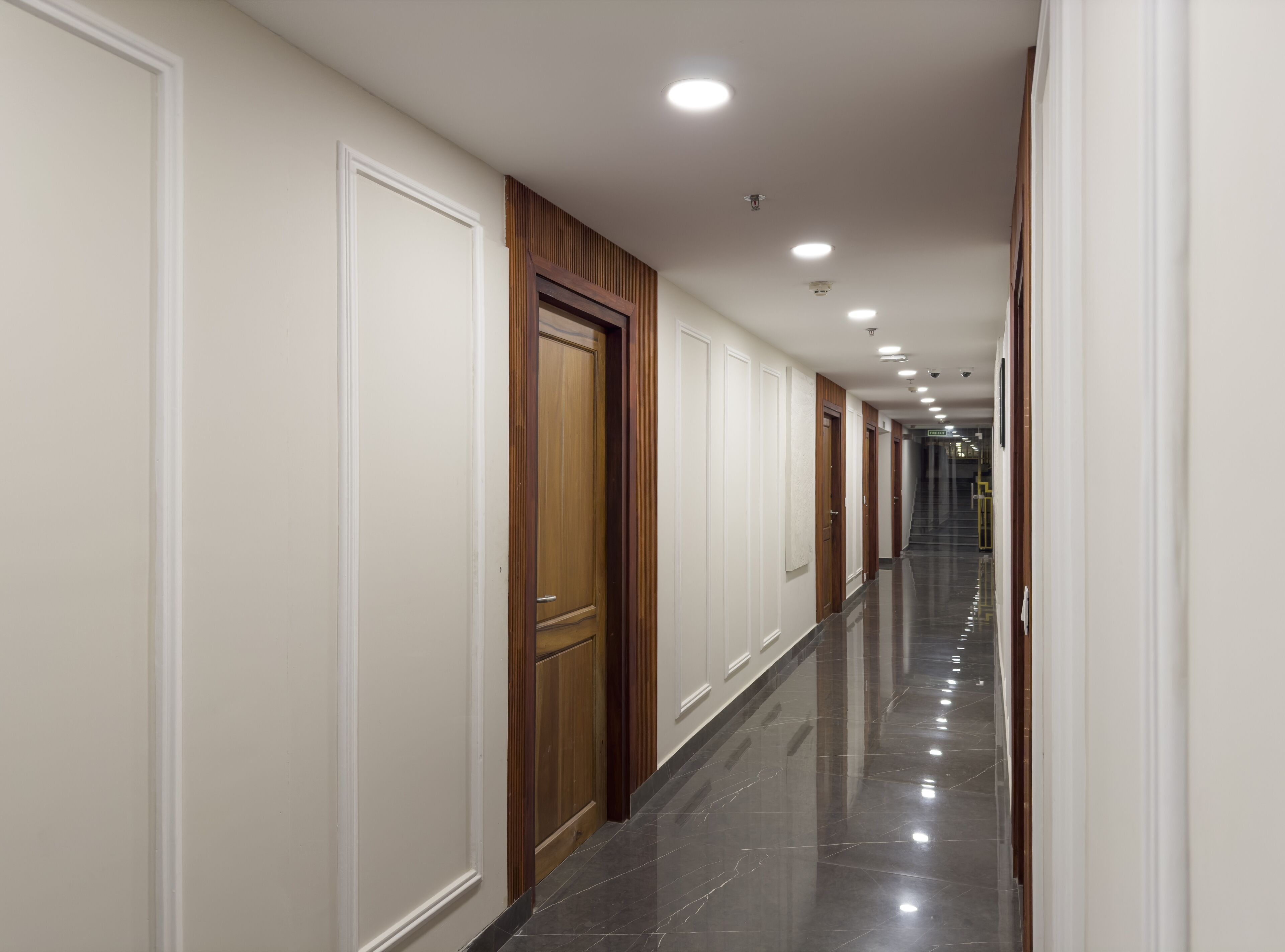 Un pasillo largo y estrecho con paredes blancas, puertas de madera y un piso de baldosas negras brillantes.