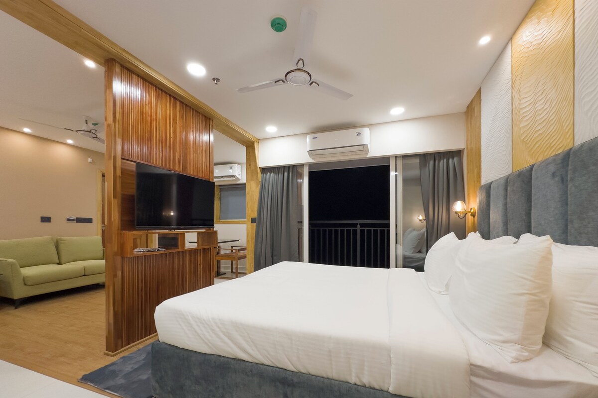 A modern hotel room with a large bed, a television, a ceiling fan, and a balcony with a view.