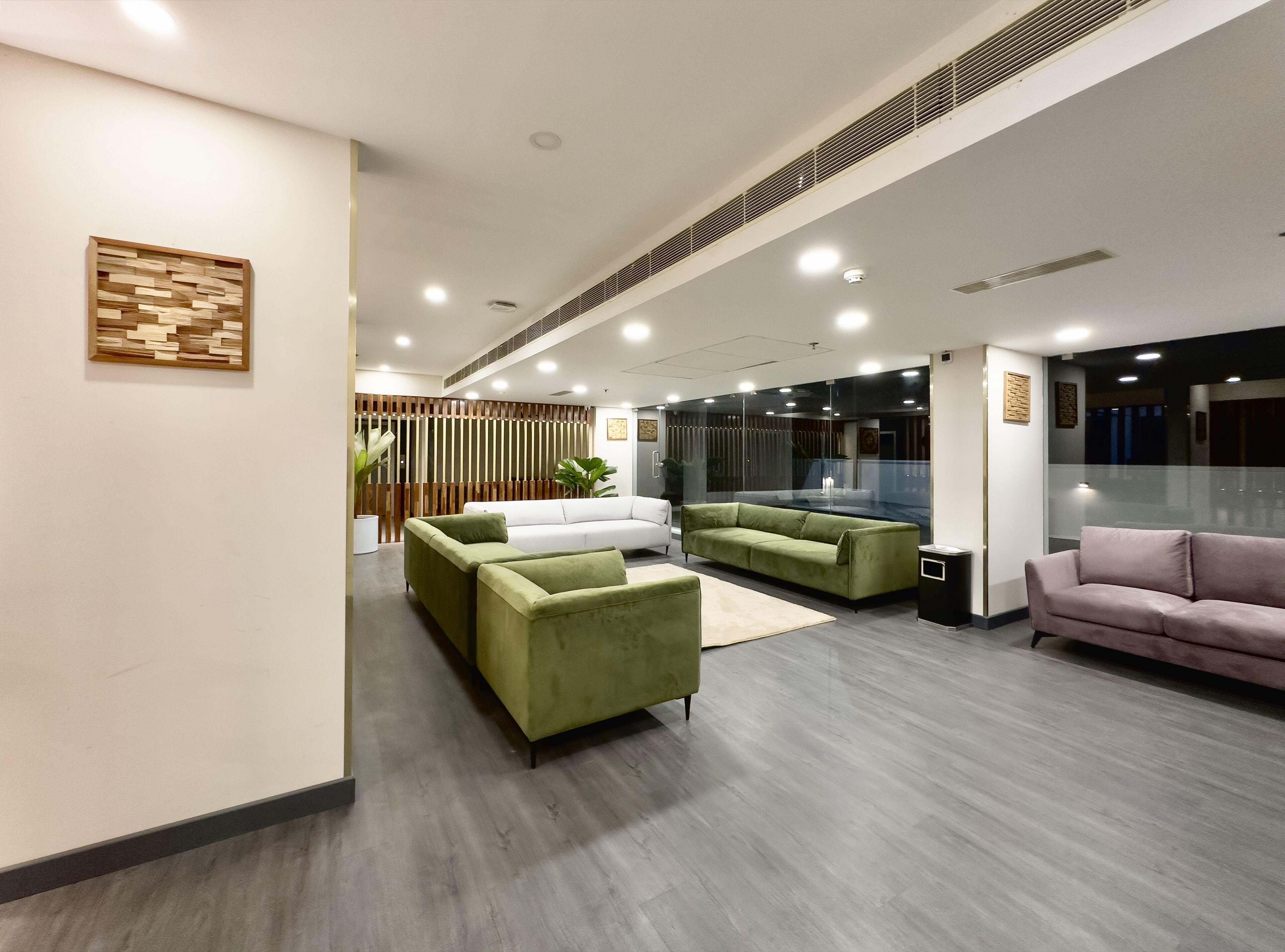 Un vestíbulo moderno con un sofá verde, un sofá blanco y un sofá rosa. Hay una partición de madera con listones verticales y una planta en maceta.