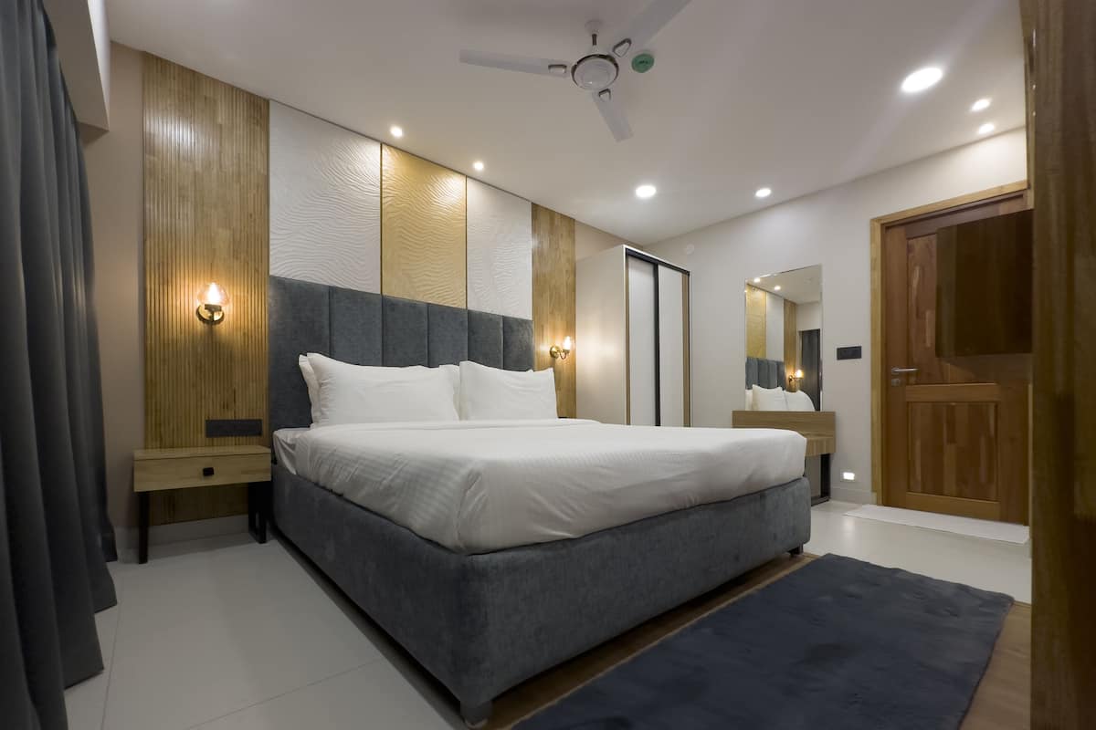 A modern bedroom with a large bed, wooden panel walls, recessed ceiling lights, a ceiling fan, and a wooden door.