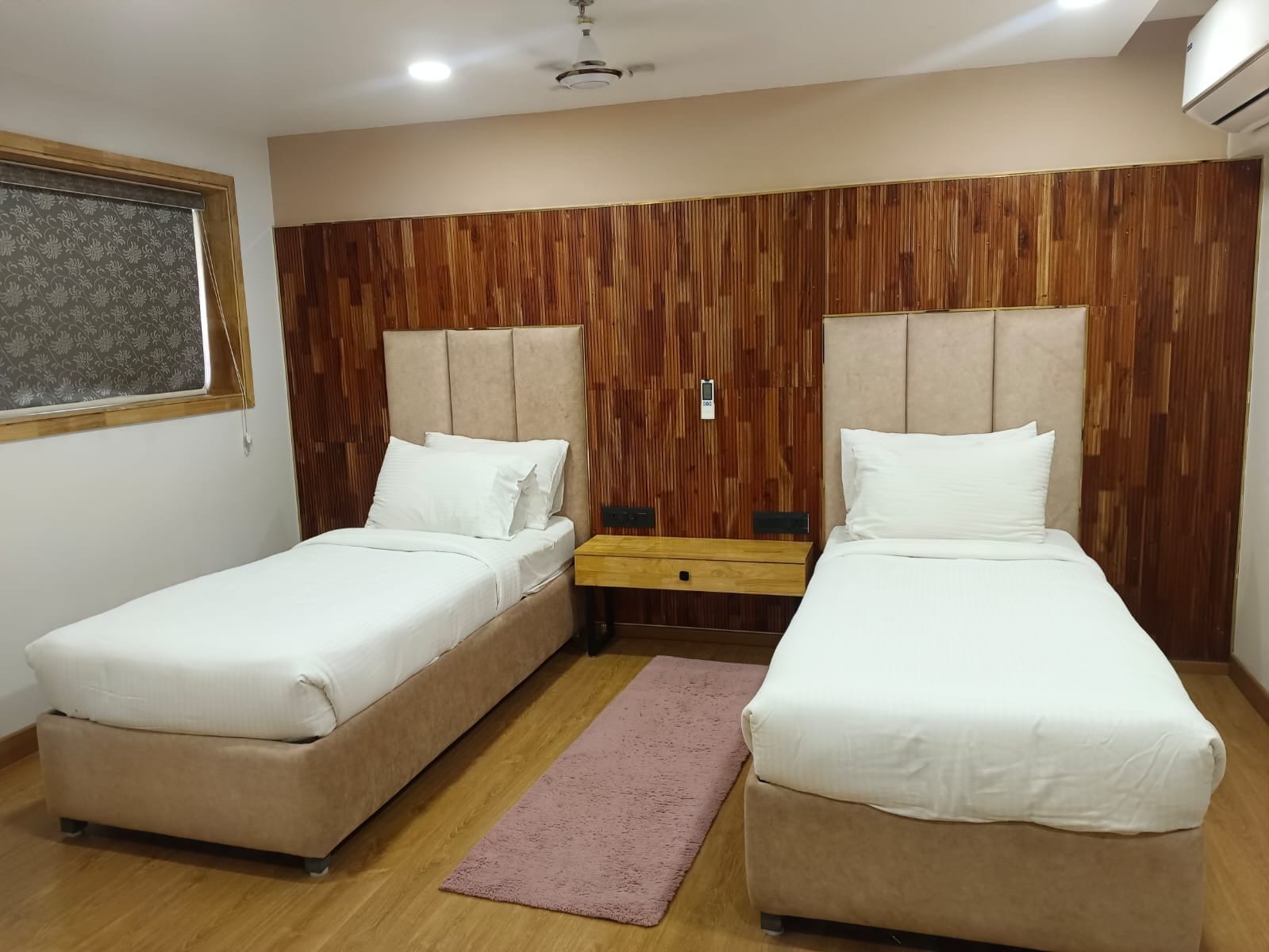 A hotel room with two beds, a wooden headboard, a small wooden nightstand, and a window with a patterned blind.