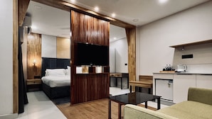 A modern hotel room with a bed, a desk, a chair, a TV, and a kitchenette. - The Habitat Suites (Chavakkad)