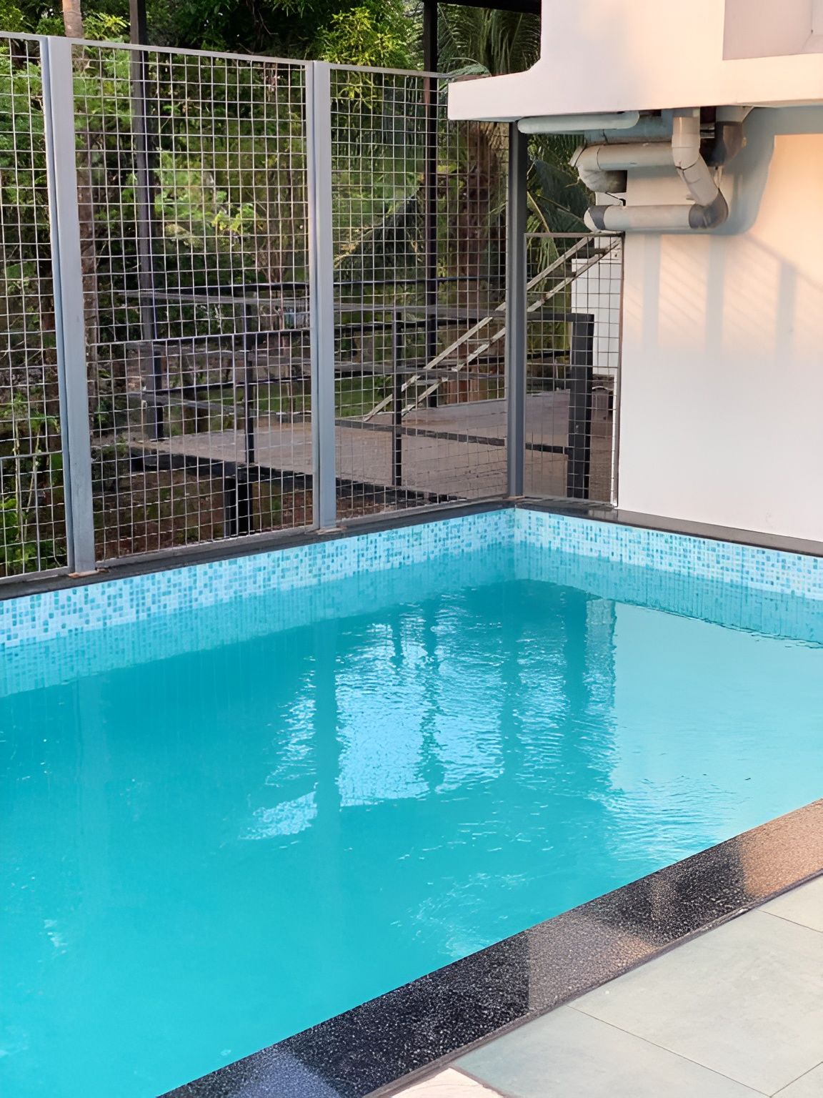 Une piscine dont le bord est revêtu de carreaux, entourée d’une clôture en métal et à laquelle on accède par un escalier menant à un niveau supérieur.