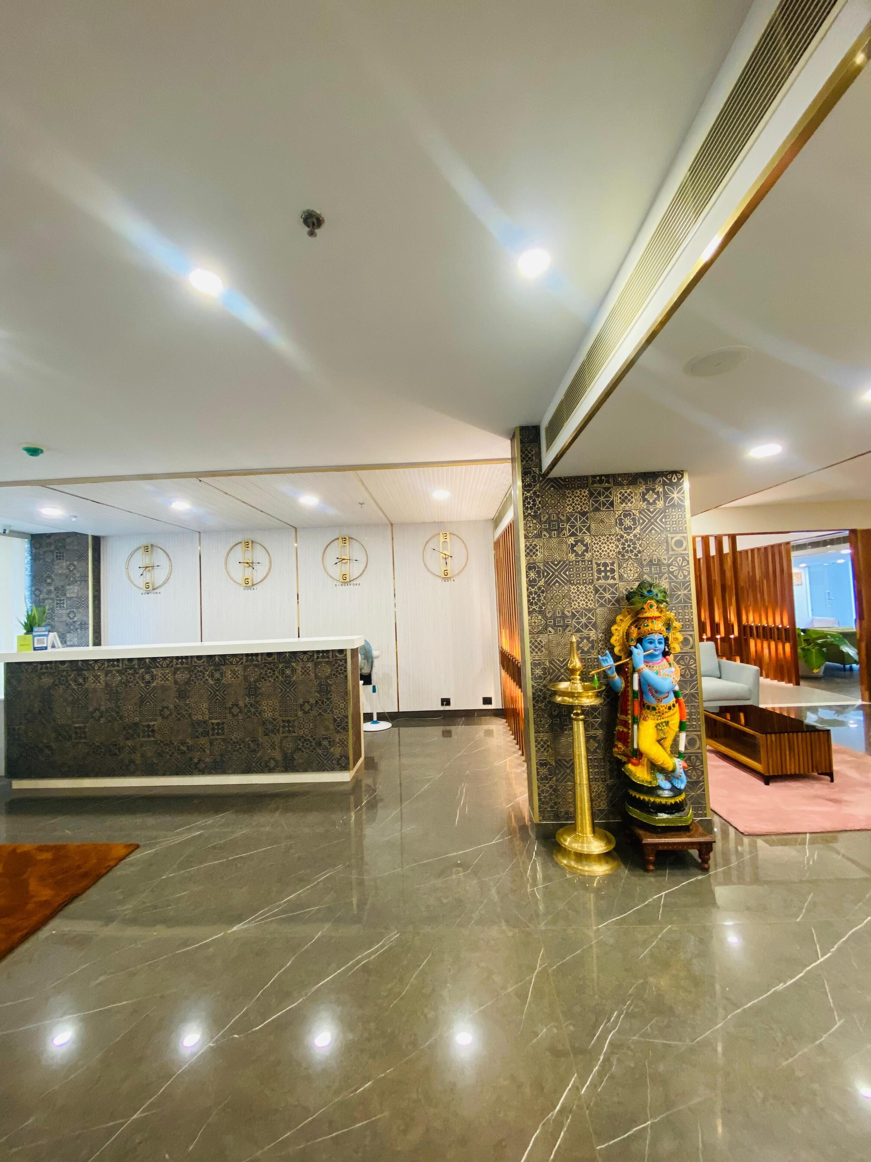 Un hall d’hôtel moderne avec un comptoir d’accueil, des horloges murales et une statue représentant une divinité.