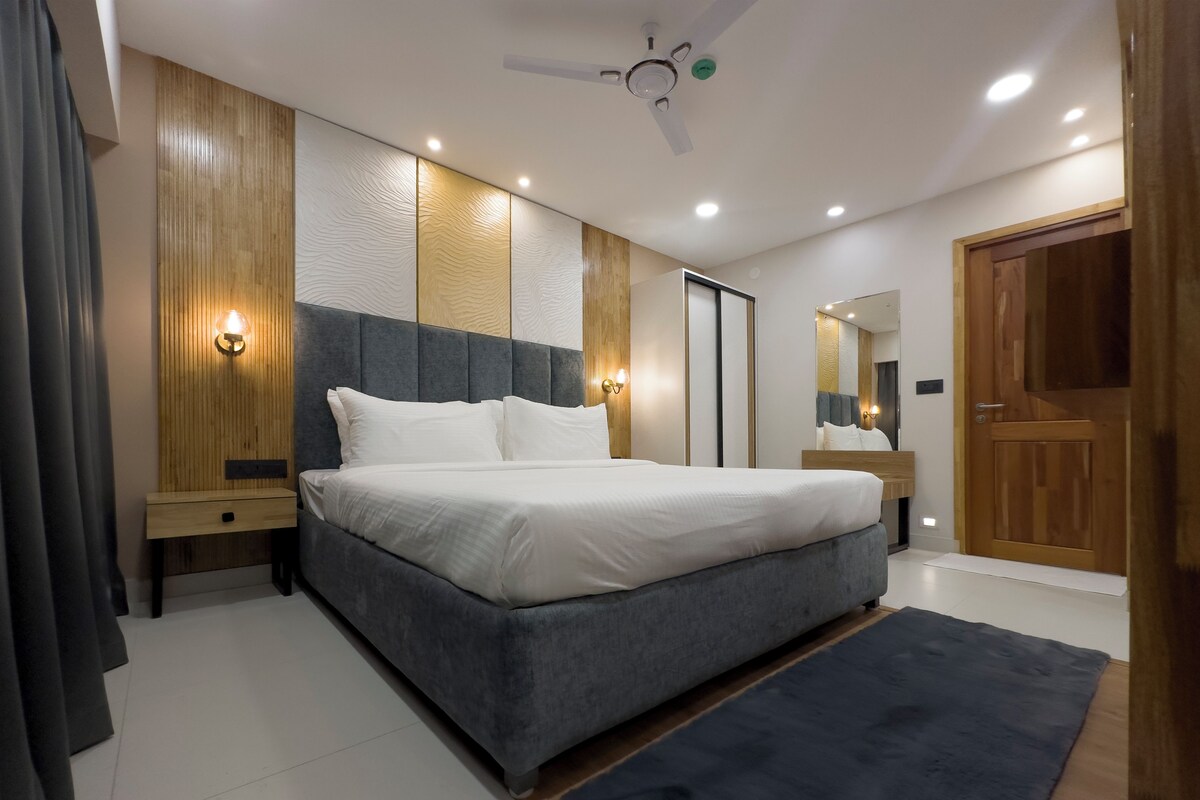 A modern bedroom with a large bed, wooden panel walls, recessed ceiling lights, a ceiling fan, and a wooden door.