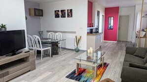 Uma sala de estar moderna com uma mesa de centro de vidro, um sofá e uma TV. Há uma área de jantar com cadeiras brancas e uma mesa de vidro. A cozinha tem uma parede com detalhes vermelhos e eletrodomésticos modernos.