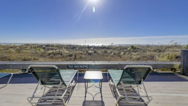 Een zonnig buitenterrein met metalen stoelen en een tafel, een 'NO SMOKING' bord, en uitzicht op het strand en de duinen.