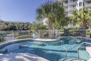 A swimming pool with a white fence, surrounded by palm trees and a multi-story building in the background.