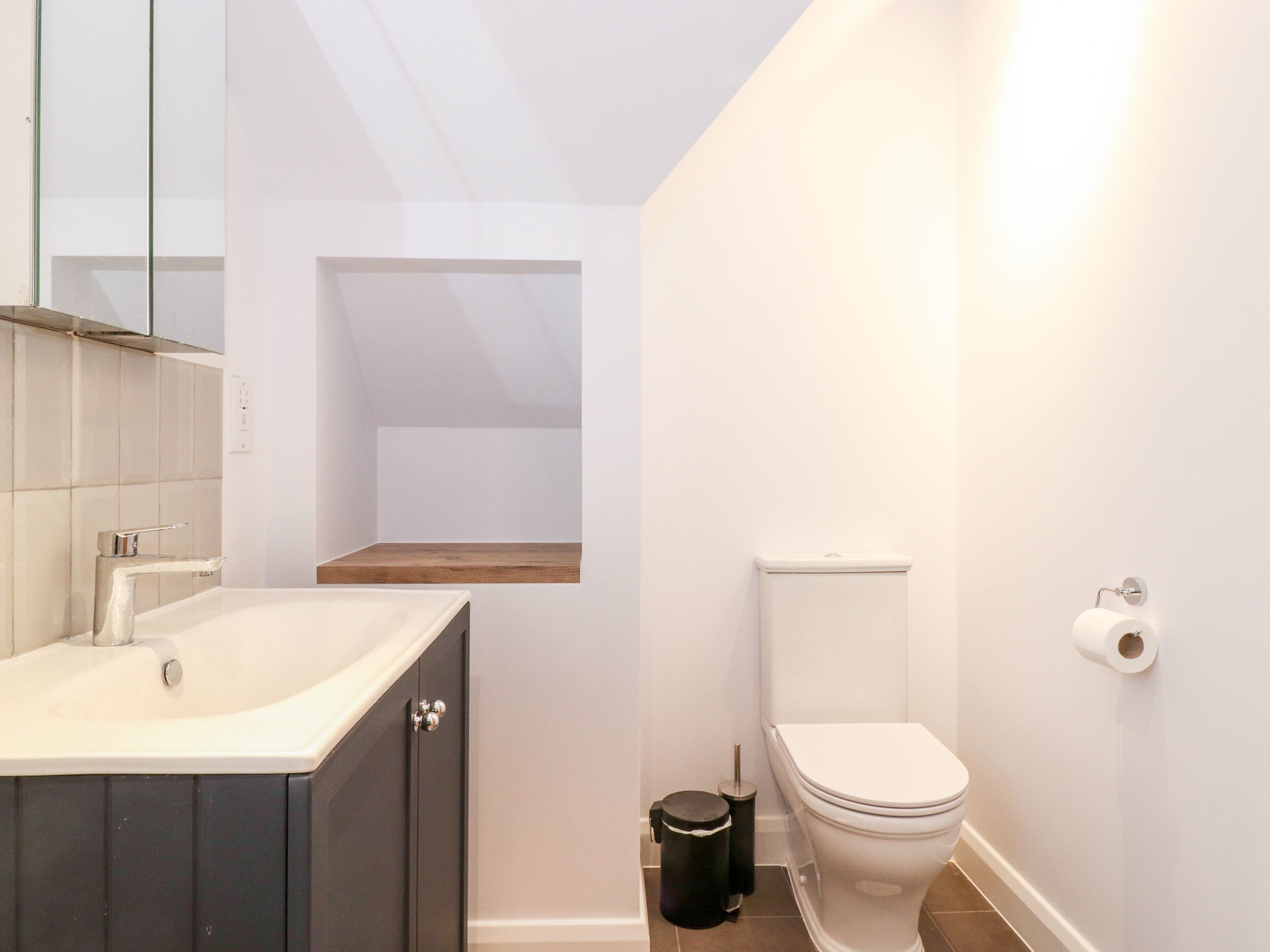 A modern bathroom with a white sink, dark cabinets, a toilet, and a small window.