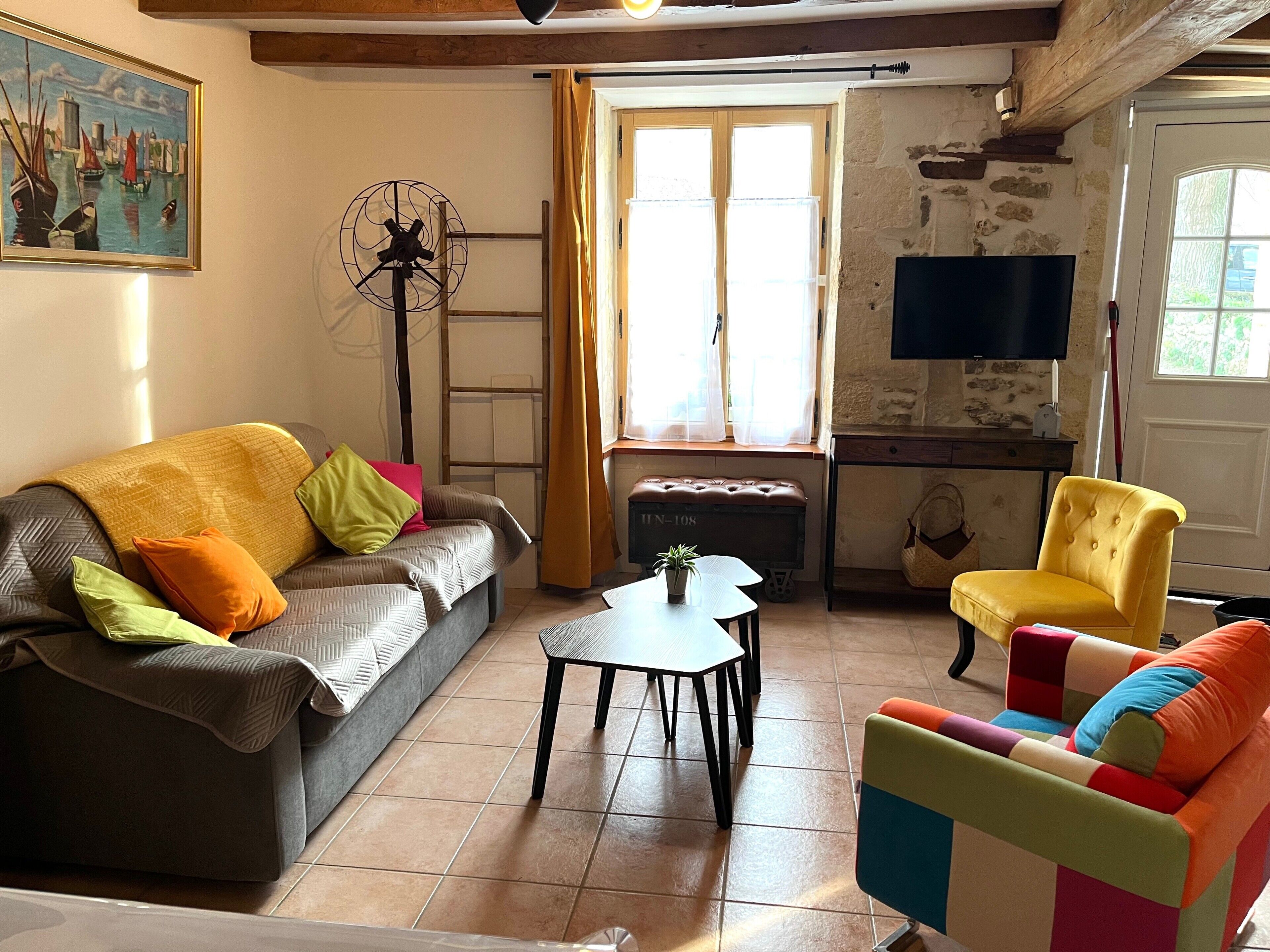 Een woonkamer met een bank, een kleurrijke fauteuil, een bijzettafeltje, een ventilator, een televisie en een raam met gordijnen.