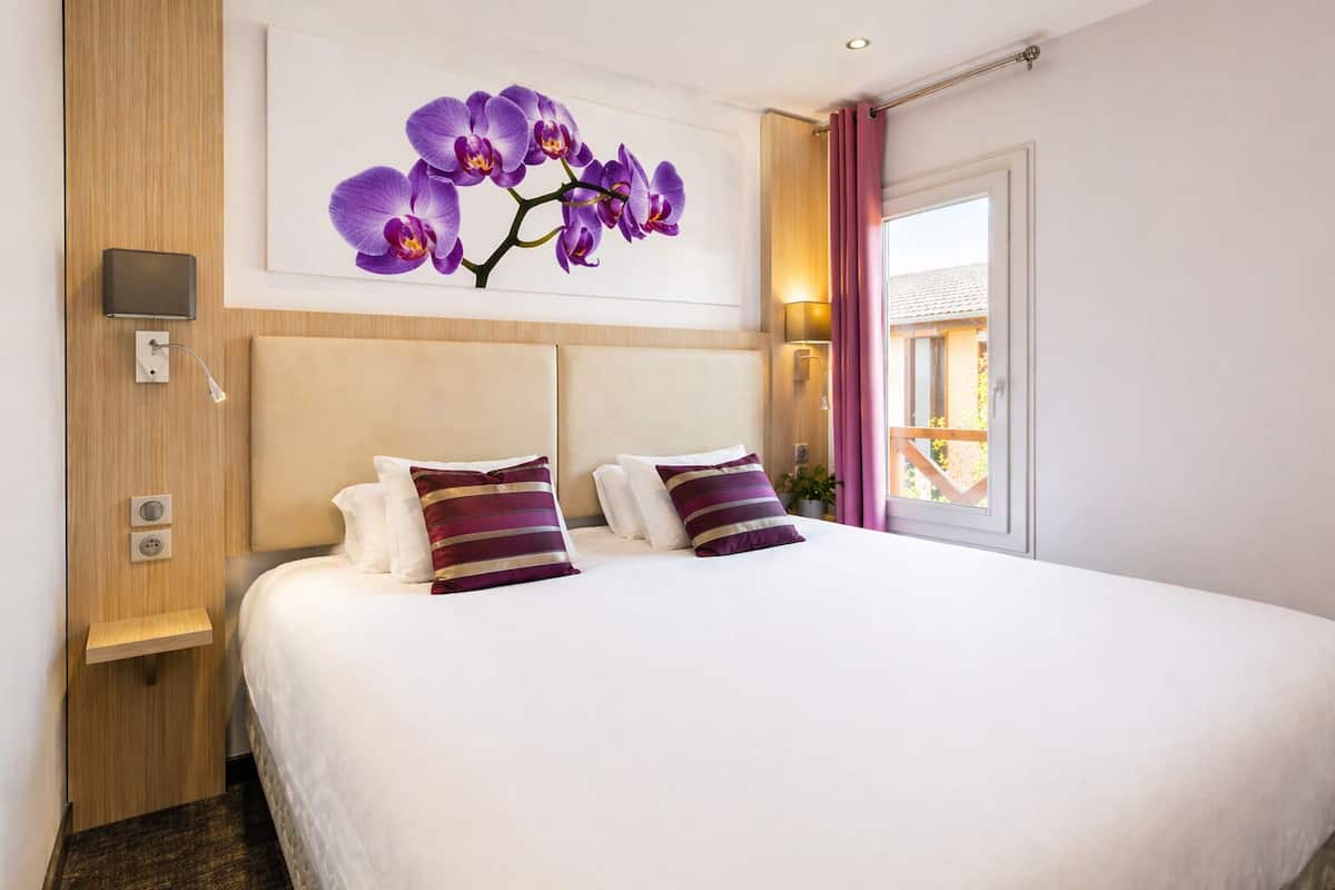 Ett hotellrum med en stor säng, en sänggavel, en väggmonterad lampa, ett eluttag och ett fönster med gardiner.