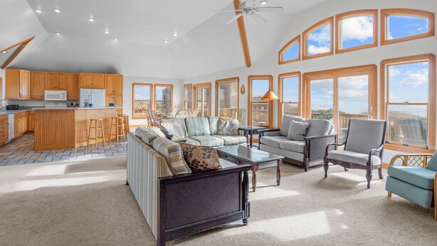 A spacious living area with a large sofa, a coffee table, and a dining table. There is a kitchen area with wooden cabinets and a microwave. The room has large windows with a view of the outdoors. - Oceanfront w/ Pool, Hot Tub, Elevator, Game Room. Feel like an Island Princess! (Frisco)